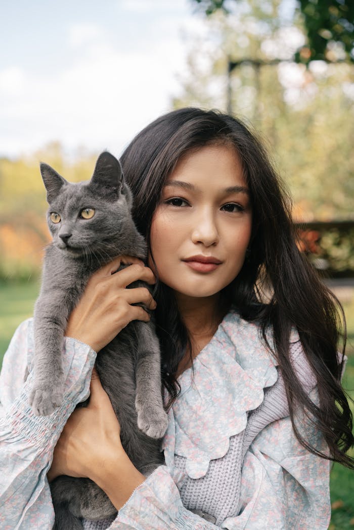 A young woman holds a gray cat outdoors in a serene setting, showcasing companionship and beauty.